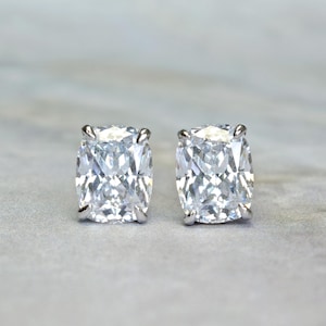 May include: A pair of cushion-cut diamond stud earrings. Each earring features a large, square-shaped, clear gemstone set in a silver metal setting. The earrings are displayed on a light gray surface.