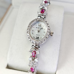 Può includere: Un orologio da polso argentato con un quadrante ovale e un cinturino decorato con pietre preziose chiare e rosse. Il quadrante presenta numeri romani e le parole "Royal Crown Jewelry". Il cinturino è composto da gruppi alternati di pietre chiare e rosse.