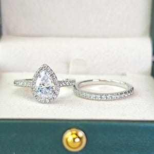 Puede incluir: Un conjunto de anillos de compromiso de plata. El anillo principal presenta una piedra central en forma de pera rodeada por un halo de pequeños diamantes. El conjunto se presenta en una caja de joyería verde azulado con un cierre dorado.