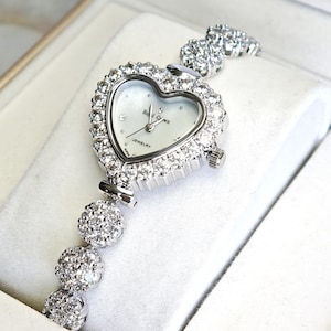 Puede incluir: Un reloj de pulsera plateado con esfera en forma de corazón, incrustado con cristales brillantes. La esfera es blanca y tiene el texto "Royal Crown Jewelry". La correa presenta esferas con cristales. Una pieza de joyería lujosa.