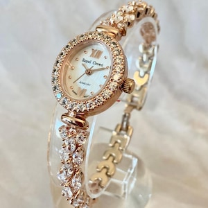 Puede incluir: Reloj de pulsera en tono oro rosa con esfera blanca y detalles de cristal. La esfera del reloj tiene el texto "Royal Crown Jewelry". La correa presenta racimos de cristal y un delicado diseño de eslabones.