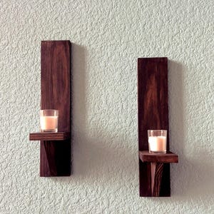 Op de afbeelding: Twee donker gebeitste houten wandkandelaars, elk met een klein plankje met een brandende kaars in een glazen votief. De kandelaars zijn bevestigd aan een getextureerde, lichtblauwe muur.