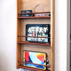 May include: Wooden wall-mounted charging station with three shelves. The top shelf holds a small digital clock and glasses. The middle shelf displays a tablet showing a video. The bottom shelf holds a tablet and a charging cable.