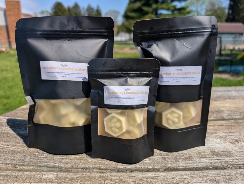 May include: Three black resealable bags filled with white wax melts shaped like honeycombs. The bags have labels that say "Strawberry Lemonade Melts".