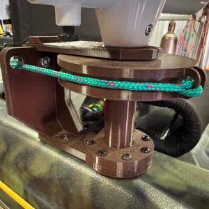 May include: A brown kayak motor mount with a white motor base and a green and red rope. The mount is attached to a camouflage kayak. The mount has a circular base with several screws.