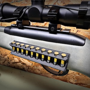 May include: A rifle with a black scope and a black magazine holder filled with yellow and brass bullets. The rifle has a green-gray stock and a black barrel. The image is taken from a close-up angle.
