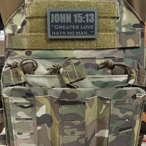 May include: A camouflage tactical vest with a black and green patch that reads "JOHN 15:13 "GREATER LOVE HATH NO MAN..."