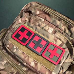 May include: Three red and black medical cross patches attached to a camouflage backpack.