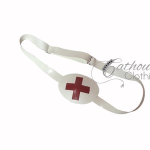 May include: White eye patch featuring a red cross, attached to a white strap with a metal clasp. The eye patch is designed to cover one eye. The text "Cathouse Clothing" is visible in the background.