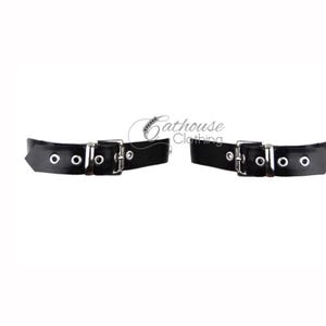 Puede incluir: Dos esposas de cuero negro con hebillas y ojales plateados. Cada esposa tiene tres ojales y una hebilla rectangular. Las esposas son de Cathouse Clothing.