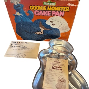 May include: A vintage Cookie Monster cake pan set. The set includes a metal Cookie Monster shaped pan, a box with a Cookie Monster illustration, and instructions. The box is orange and features the Sesame Street logo.