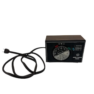 May include: A GE Home Sentry timer with a dark wood-grain finish housing and a black control panel. The circular dial features AM and PM markings, daytime and nighttime zones, and red and green setting pins. A black power cord is attached.