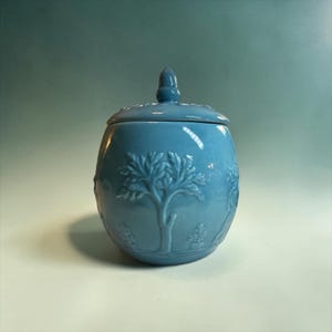 May include: A light blue ceramic jar with a matching lid featuring a finial handle. The exterior surface is decorated with embossed relief designs of trees and foliage, creating a textured pattern around the rounded body of the container.