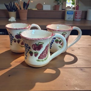 May include: Three ceramic mugs with white handles and a cream-colored base. Each mug is decorated with hand-painted red berries, green leaves, and blue grapes. The rims are painted red. The mugs are arranged on a wooden surface.