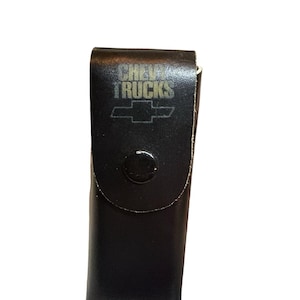 May include: A black leather belt pouch featuring a snap button closure. The front of the pouch is embossed with the text CHEVY TRUCKS and the Chevrolet bowtie logo. The rectangular case has a smooth finish and a vertical orientation.