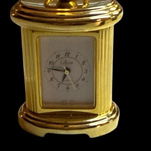 May include: A small gold-toned metal carriage clock with a rectangular white face featuring black numerals and the text "Colleria". The clock has a fluted column design on the sides, a smooth base, and a decorative handle on the top.