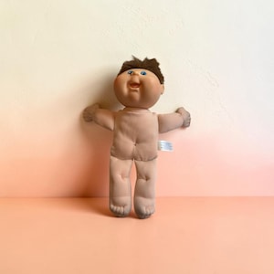 May include: A doll with a soft, light-toned fabric body, a molded vinyl head, and short brown hair. The doll features blue eyes, a small open mouth, and outstretched arms. A small white fabric tag is visible on the side of the doll's torso.