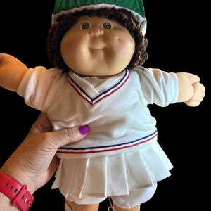 May include: A doll with brown yarn hair wearing a green visor, a white V-neck shirt with red and blue trim, a white pleated skirt, and white sneakers with blue stripes. The doll has a round face, dimples, and a light skin tone.