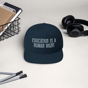 May include: A navy blue baseball cap with the text "EDUCATION IS A HUMAN RIGHT" embroidered in white on the front.