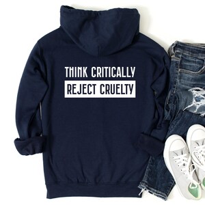 May include: A navy blue hoodie with the text "THINK CRITICALLY REJECT CRUELTY" in white. The hoodie is paired with blue jeans and gray sneakers on a white background. The hoodie has a hood and long sleeves.