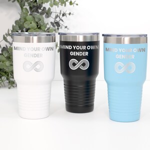 Może przedstawiać: Trzy kubki ze stali nierdzewnej z grawerowanym napisem "Mind Your Own Gender" i symbolem nieskończoności. Kubki są białe, czarne i jasnoniebieskie.