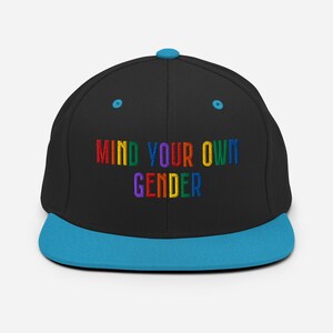 May include: A black and blue baseball cap with the text "MIND YOUR OWN GENDER" embroidered in rainbow colors.