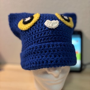 Puede incluir: Un gorro azul tejido a mano con ojos de gato amarillos y negros y una nariz blanca. El gorro se exhibe en una cabeza de maniquí blanca. El gorro es un accesorio único y caprichoso.