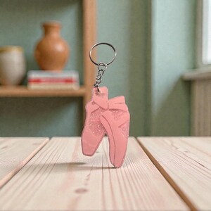 May include: A pink ballet slipper keychain with glitter accents. The keychain features two ballet slippers, each with a bow. The keychain is attached to a silver-colored ring and chain. The background includes a wooden surface and a shelf with decorative items.