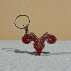 May include: Red and silver Aries zodiac sign keychain. The keychain features a stylized ram symbol with a glittery silver outline and accents. The keychain is attached to a silver metal ring.