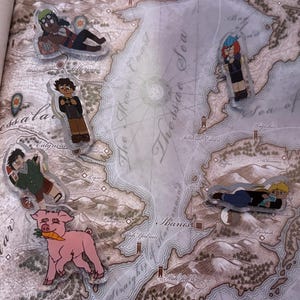 Può includere: Una collezione di spille in acrilico raffiguranti personaggi dei cartoni animati e un maiale rosa con una carota. Le spille sono posizionate su una mappa con testo. I personaggi sono in varie pose.