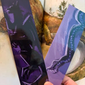 May include: Two rectangular bookmarks featuring fantasy creature illustrations. One bookmark is mainly purple with a black silhouette of a dragon-like creature. The other is light purple with a blue illustration of a creature's tail. The bookmarks are held over an open book.