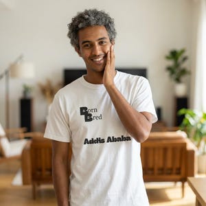 May include: White t-shirt with the text "Born Bred Addis Ababa" in black. The shirt is being worn by a person with gray curly hair. The person is smiling and touching their face. The background includes a wooden cabinet and a plant.