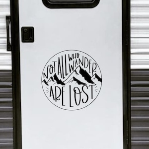 Può includere: Una porta di camper bianca con un adesivo in vinile nero che recita "Not all who wander are lost" con una grafica di una catena montuosa. L'adesivo è in un cerchio con un contorno nero. Una ghirlanda marrone è appesa alla porta.