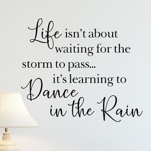 May include: A wall decal with a quote that reads "Life isn't about waiting for the storm to pass... it's learning to Dance in the Rain". The decal is on a white wall above a wooden dresser with a lamp and books on top.