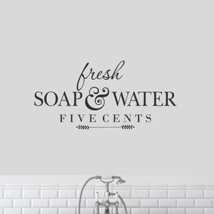 May include: A black and white bathroom sign with the text "fresh soap & water five cents" with a decorative flourish and a small arrow on each side of the text. The sign is on a white tiled wall above a black bathtub.