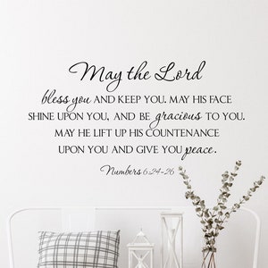 Puede incluir: Pared blanca con una cita en blanco y negro sobre la paz y las bendiciones de la Biblia, Números 6:24-26. La cita se muestra sobre un banco blanco con un cojín a cuadros, dos linternas y dos cestas de mimbre. El texto "Sugar Creek" se muestra en la esquina inferior derecha.