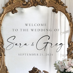 Może przedstawiać: Złota, ozdobna rama lustra z czarną naklejką winylową z napisem WELCOME TO THE WEDDING OF Sara & Greg SEPTEMBER 21, 2024. Lustro jest udekorowane białym wazonem z kwiatami oraz stosami książek u podstawy ramy.