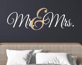 May include: A wall decal featuring the words "Mr. & Mrs." in elegant script. The word "Mr." and "Mrs." are in white, with the ampersand in a gold color. The decal is displayed above a bed with white pillows and a woven headboard.