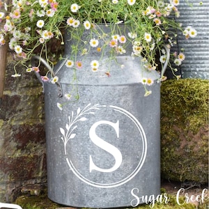 May include: A galvanized metal milk can overflowing with small white and pink daisies. The can features a white monogram "S" within a circle and a floral design. The words "Sugar Creek" are at the bottom.