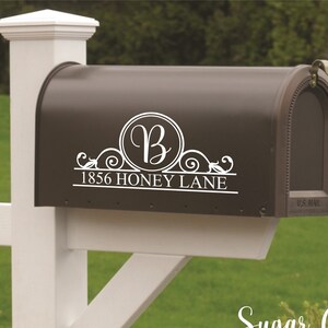 May include: A dark brown mailbox with a white decorative design featuring the letter "B" inside a circle, with the address "1856 HONEY LANE" below. The mailbox is mounted on a white post.