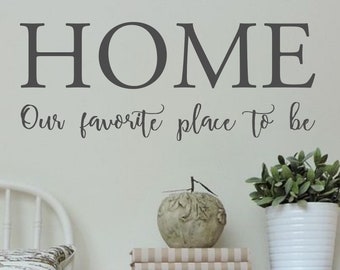 May include: A gray vinyl wall decal with the text "HOME" in a serif font and the text "Our favorite place to be" in a cursive font. The decal is displayed on a white wall above a wooden dresser with a decorative apple and books.