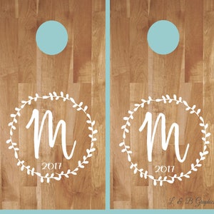 Op de afbeelding: Twee houten cornhole-borden met een wit kransontwerp en de letter "M" in het midden. De krans heeft kleine bladeren en het jaar "2017" staat onder de letter. De borden hebben een lichtblauwe cirkel in het bovenste midden van elk bord.