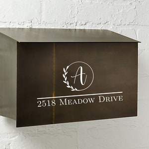 Puede incluir: Un buzón de metal marrón con un monograma blanco de la letra "A" rodeado de una corona y el texto "2518 Meadow Drive".