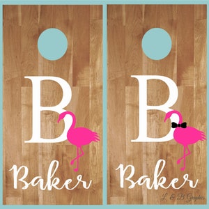 Op de afbeelding: Twee houten cornhole-borden met een witte letter B en een roze flamingo op elk. De borden hebben een blauwe cirkel in het midden en de naam "Baker" staat onder de flamingo. Het rechterbord heeft een zwarte vlinderdas op de flamingo. De rechterbenedenhoek van de afbeelding zegt "L & B Graphics".