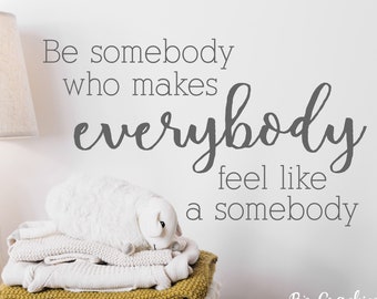 May include: A white wall with the quote "Be somebody who makes everybody feel like a somebody" written in grey text. A white lamp sits on a wooden table with a stack of white and yellow knitted blankets.
