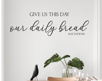 May include: A white wall with a black vinyl decal that reads "Give us this day our daily bread Matthew 6:11". The decal is above a dining table with chairs and a potted plant.