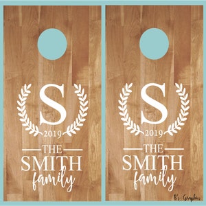 Op de afbeelding: Twee houten cornhole-borden met een lichtblauwe cirkel in het midden van elk bord. De borden zijn versierd met een witte krans, de letter "S", het jaar "2019" en de tekst "THE SMITH family".