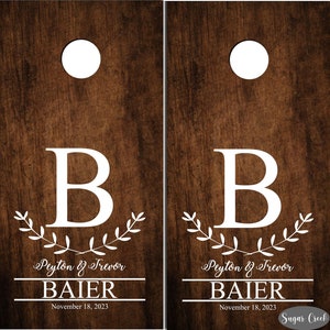 Op de afbeelding: Twee bruine houten cornhole-borden met wit geschilderde cirkels, een witte letter B, een krans van bladeren, de tekst "Peyton & Trevor", de tekst "BAIER" en de datum "18 november 2023".