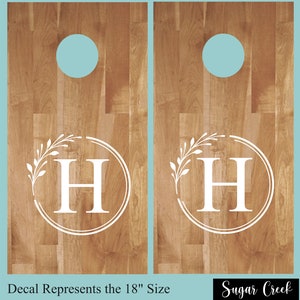 Op de afbeelding: Twee houten cornhole-borden met een witte monogram-sticker met de letter "H", omringd door een cirkel en een bladontwerp. De sticker staat voor de maat van 18 inch. De tekst "Sugar Creek" is zichtbaar onderaan de afbeelding.