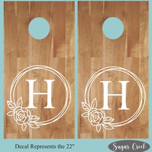 Op de afbeelding: Twee houten cornhole-borden met een witte monogram-sticker met de letter "H" in een cirkel met bloemmotieven. De sticker staat voor de maat van 22 inch. De tekst "Sugar Creek" is zichtbaar in de rechterbenedenhoek.